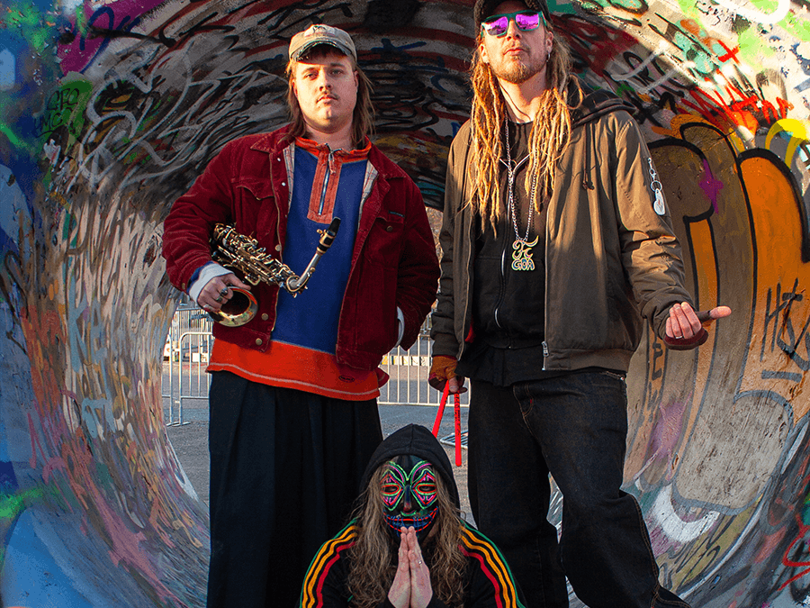 The Psy Boys trio in a graffiti tunnel, one holding a saxophone and one kneeling in a mask