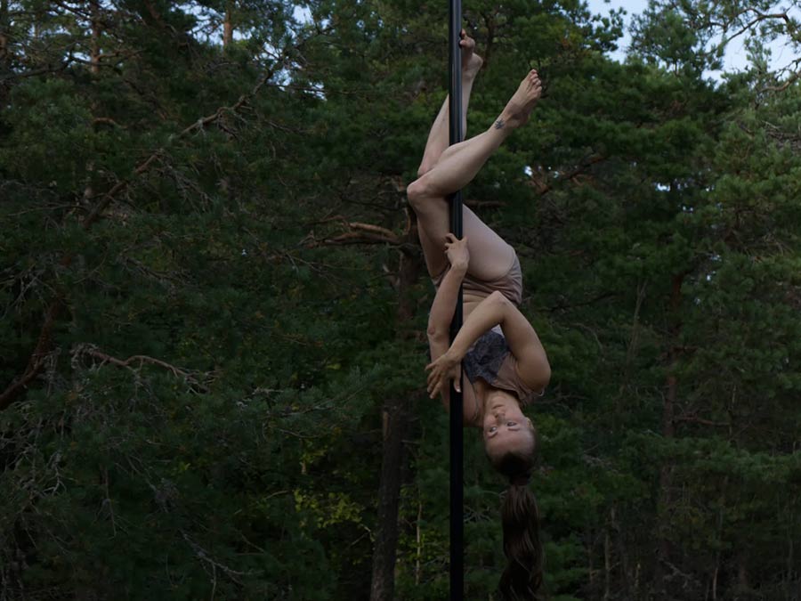 Noora Juppi upside down on a dance pole in front of pine trees