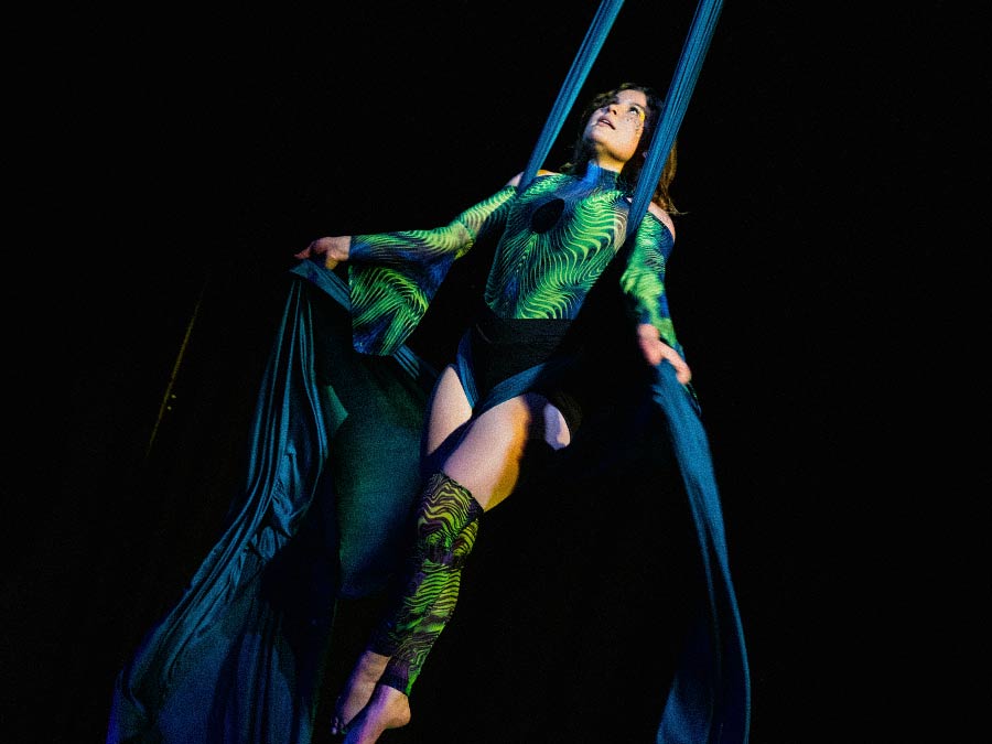 Nyx in a green bodysuit hanging in dark aerial silks