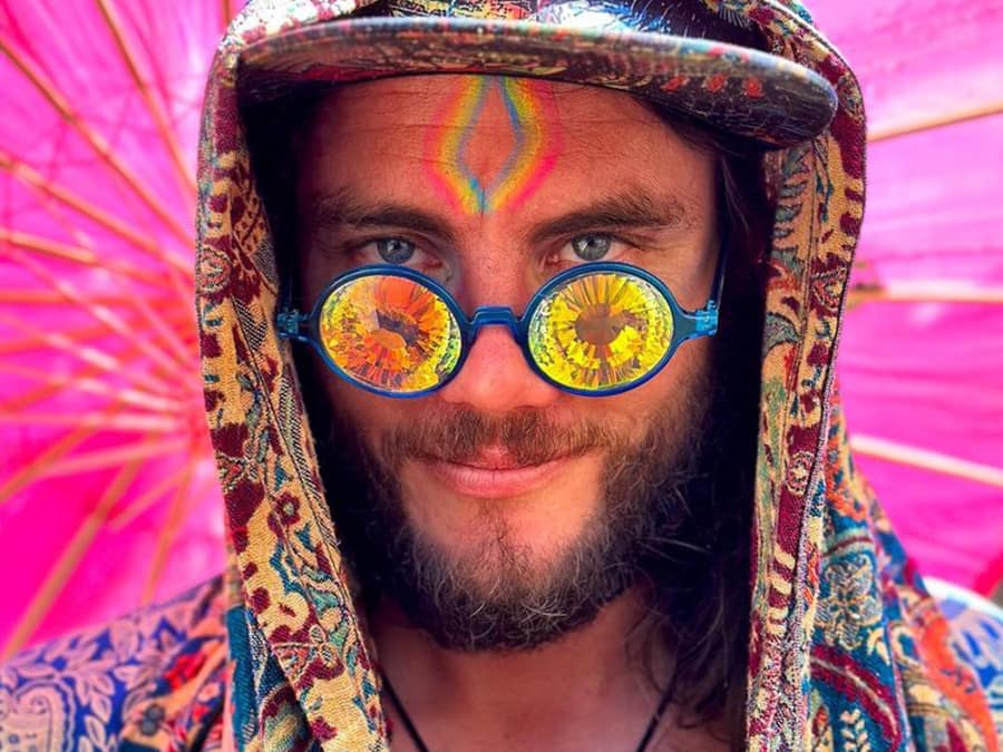 Grouch in a patterned hood, face paint and kaleidoscope glasses, smiling in front of a pink backdrop