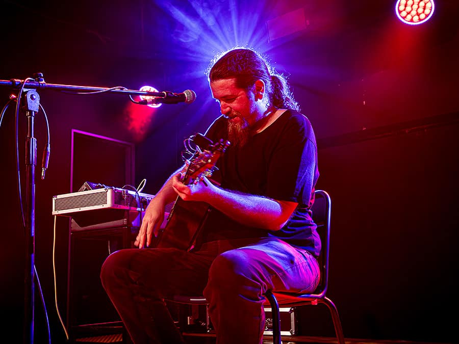 Mark Hillman playing acoustic guitar on stage under red and blue lights