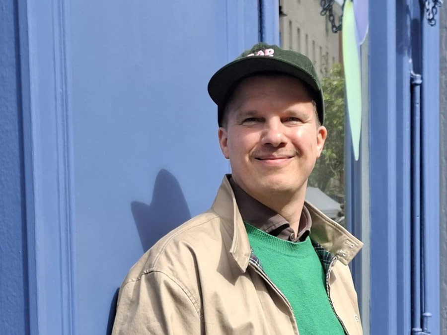 James Reipas in a cap and beige jacket, smiling against a blue wall