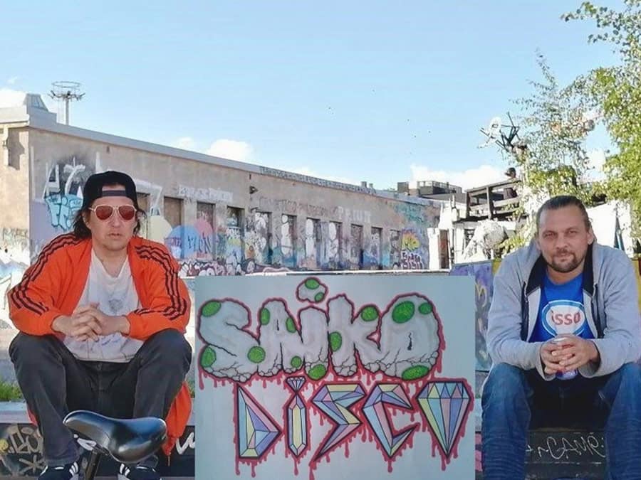 The two members of Saiko Disco sitting beside a Saiko Disco graffiti sign