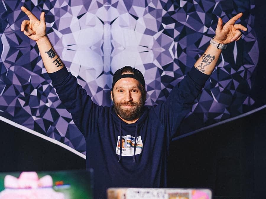 D.D.A. raising both arms behind the decks in front of a geometric backdrop
