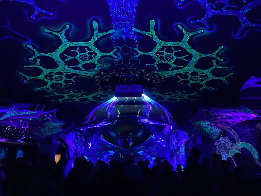 A patterned mesh canopy by Ihti Anderson glowing green and blue under UV light above a stage