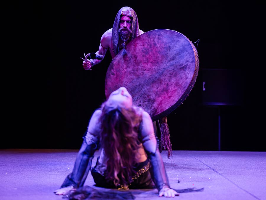 A drummer with a large frame drum standing over a dancer in a deep backbend on stage