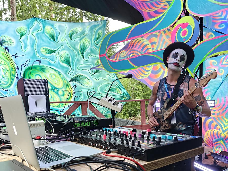 A Jazuur Space Club musician in clown make-up and a top hat playing guitar beside a laptop and mixer