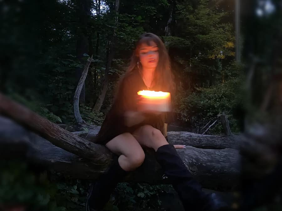 Idamine sitting on a fallen tree in a dark forest, holding a glowing light