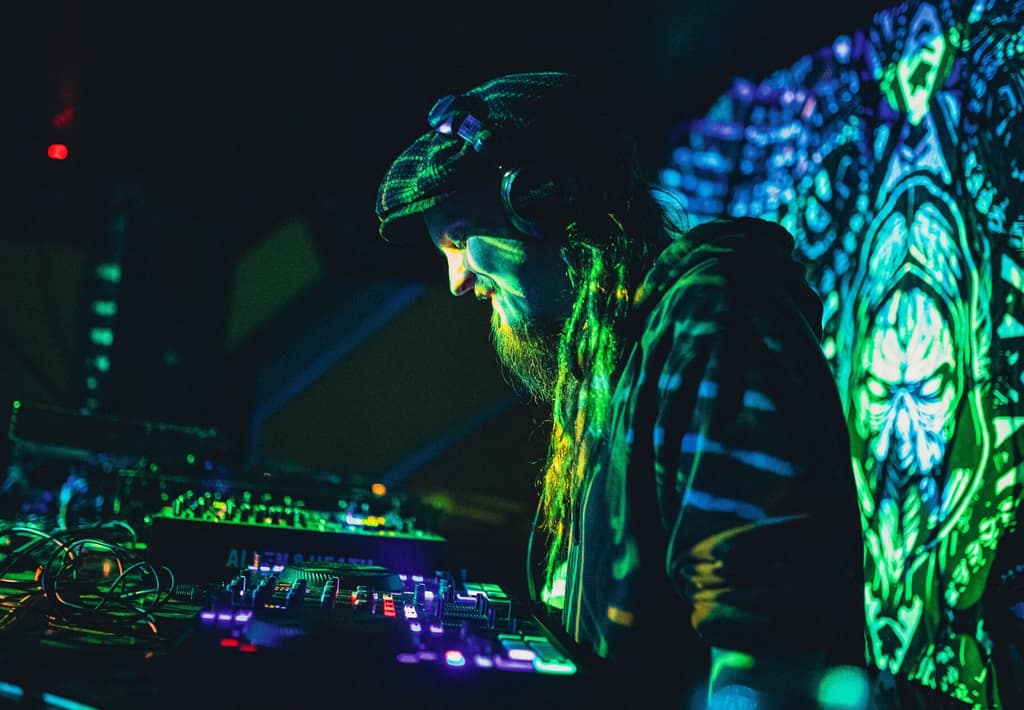 Shroomi at the controls bathed in green UV light before a psychedelic backdrop