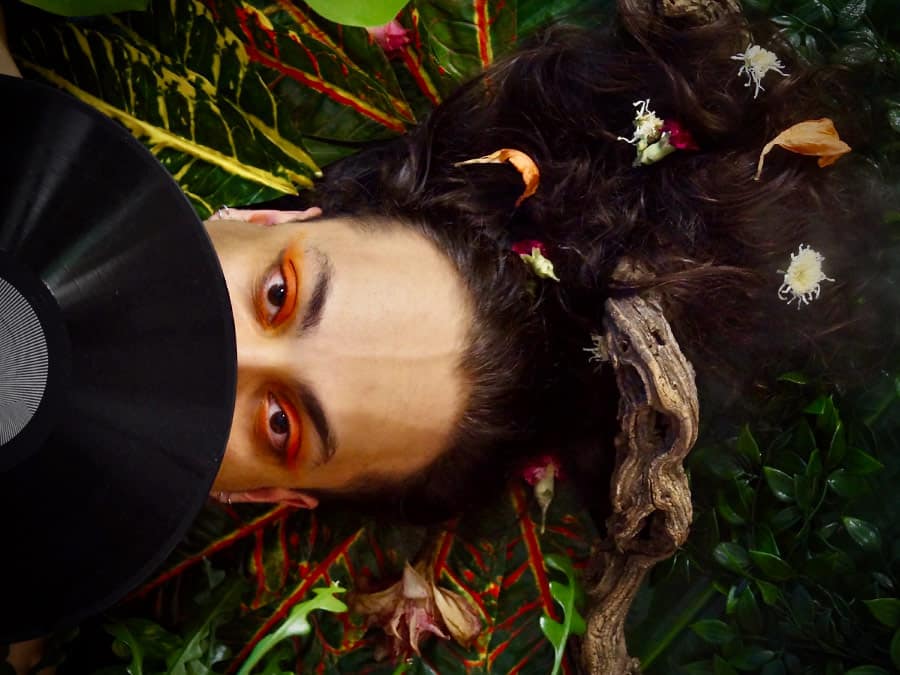 Samir Hani with orange-painted eyes lying among leaves and flowers beside a vinyl record