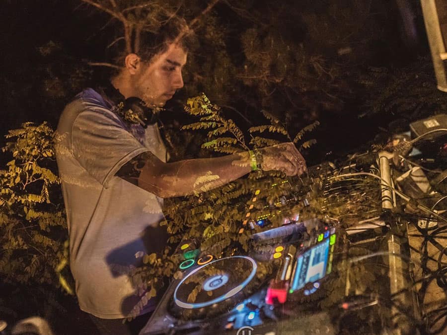 Bent playing at DJ decks outdoors at night among foliage