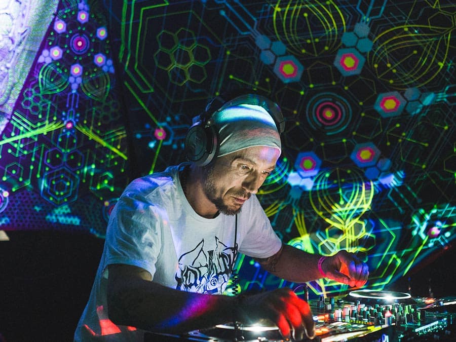 Dj Hugadam in headphones at the mixer under UV-lit psychedelic decor